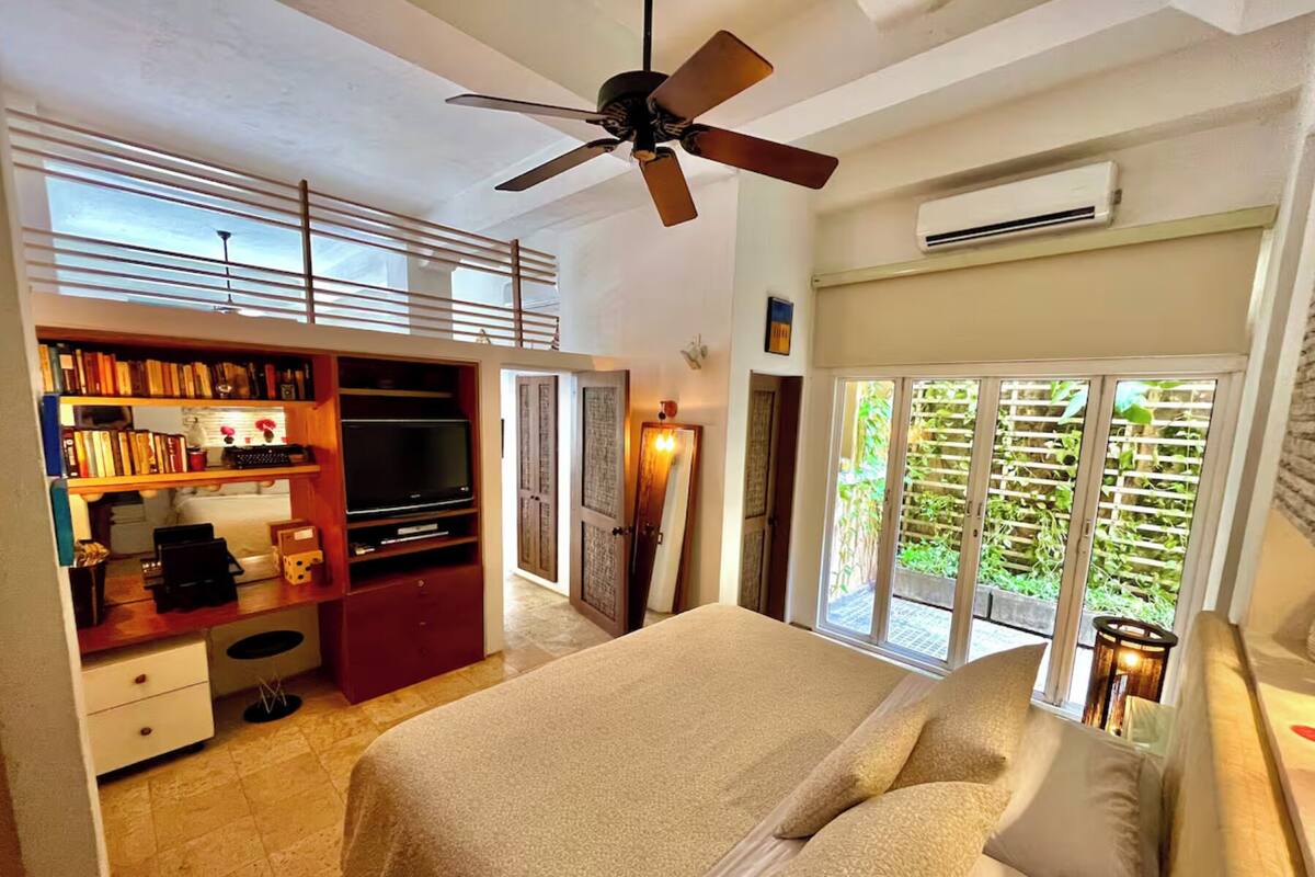 A bedroom with a bed, a ceiling fan, a large window with a view of greenery, and a built-in TV stand.