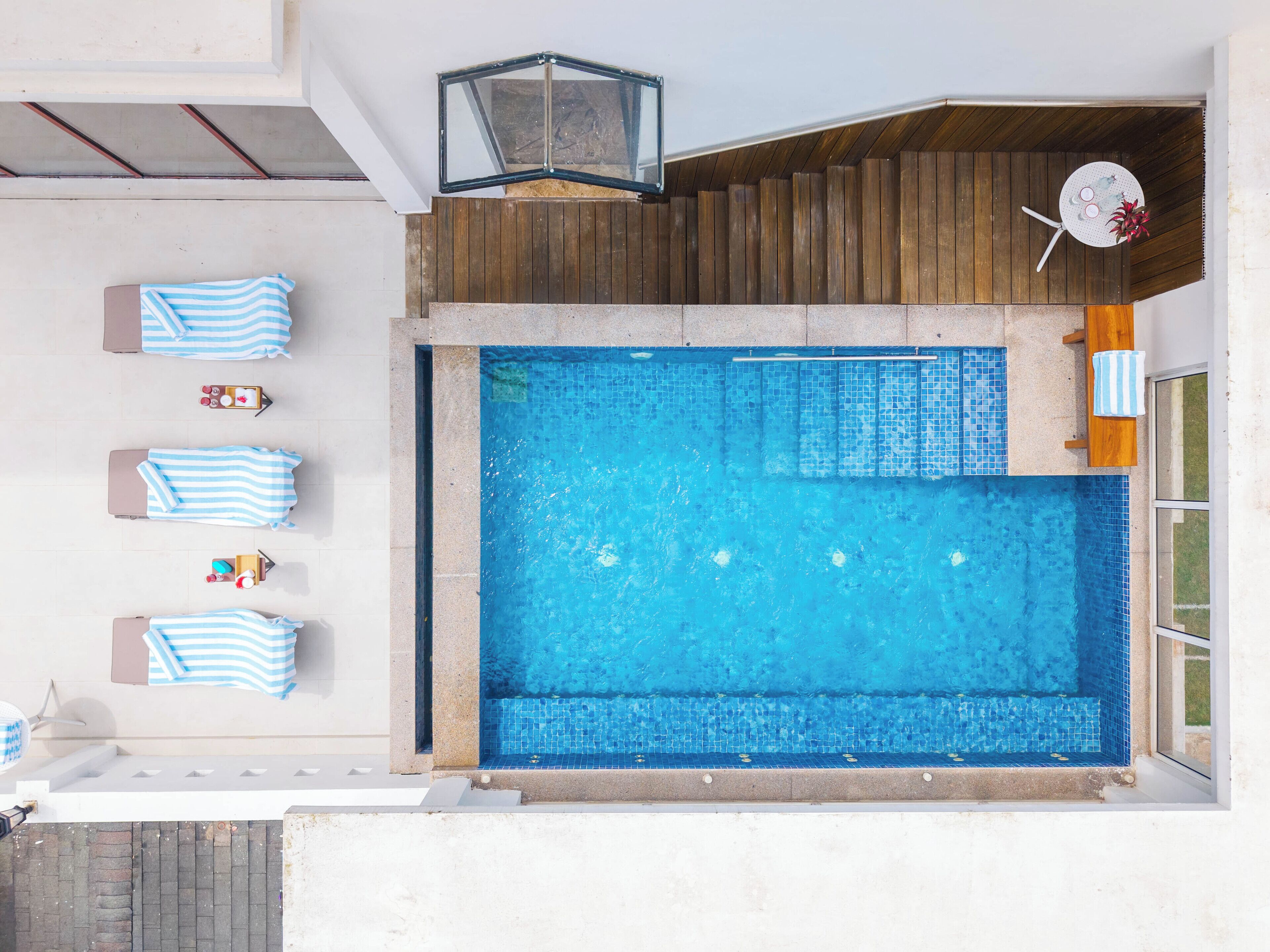 Ein Pool auf dem Dach mit einer Holzterrasse, einer Lounge-Ecke mit gestreiften Handtüchern und einem Tisch mit Getränken.