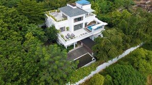 Ett modernt hus med flera våningar, omgivet av tät grönska, med en takterrass och en swimmingpool.