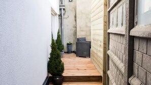 A narrow wooden pathway leading to a door, flanked by white walls and a brick wall.