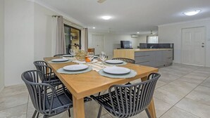A dining area with a wooden table set for six, surrounded by black chairs. A ceiling fan is above, and a kitchen area is visible in the background.