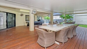 An outdoor covered patio with a wooden deck, a dining table with wicker chairs, and a pool table in the background.