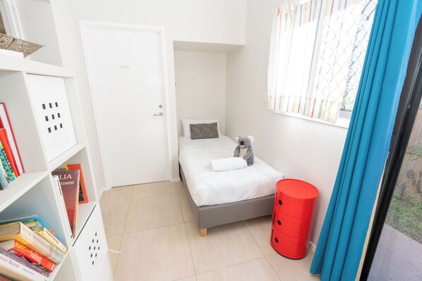 A small, neatly arranged bedroom with a bed, a red trash bin, a bookshelf with books, and a window with curtains.