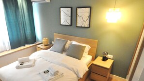 A neatly made bed with white linens and towels, a bedside table with a lamp, and two framed artworks on the wall. - Zennova 71 Mondè Thessaloniki Apt (Thessaloniki)