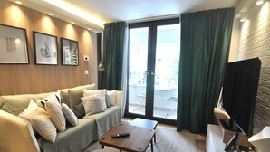 A modern living room with a sofa, a coffee table, and a TV. There are framed pictures on the wall and a view of the outside through the window. - Zennova 71 Mondè Thessaloniki Apt (Thessaloniki)