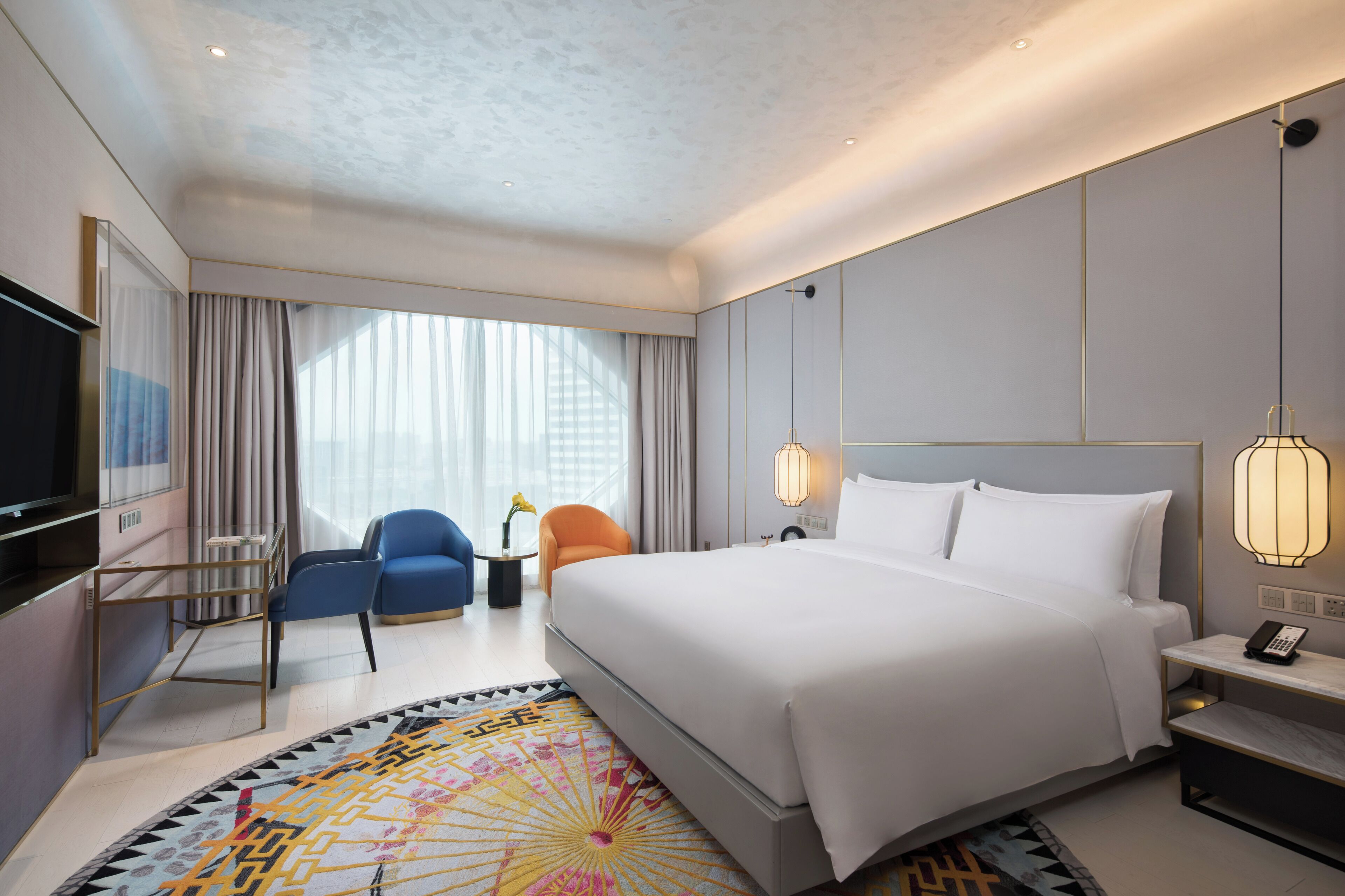 A modern hotel room with a large bed, a TV, a desk with a lamp, and a chair.