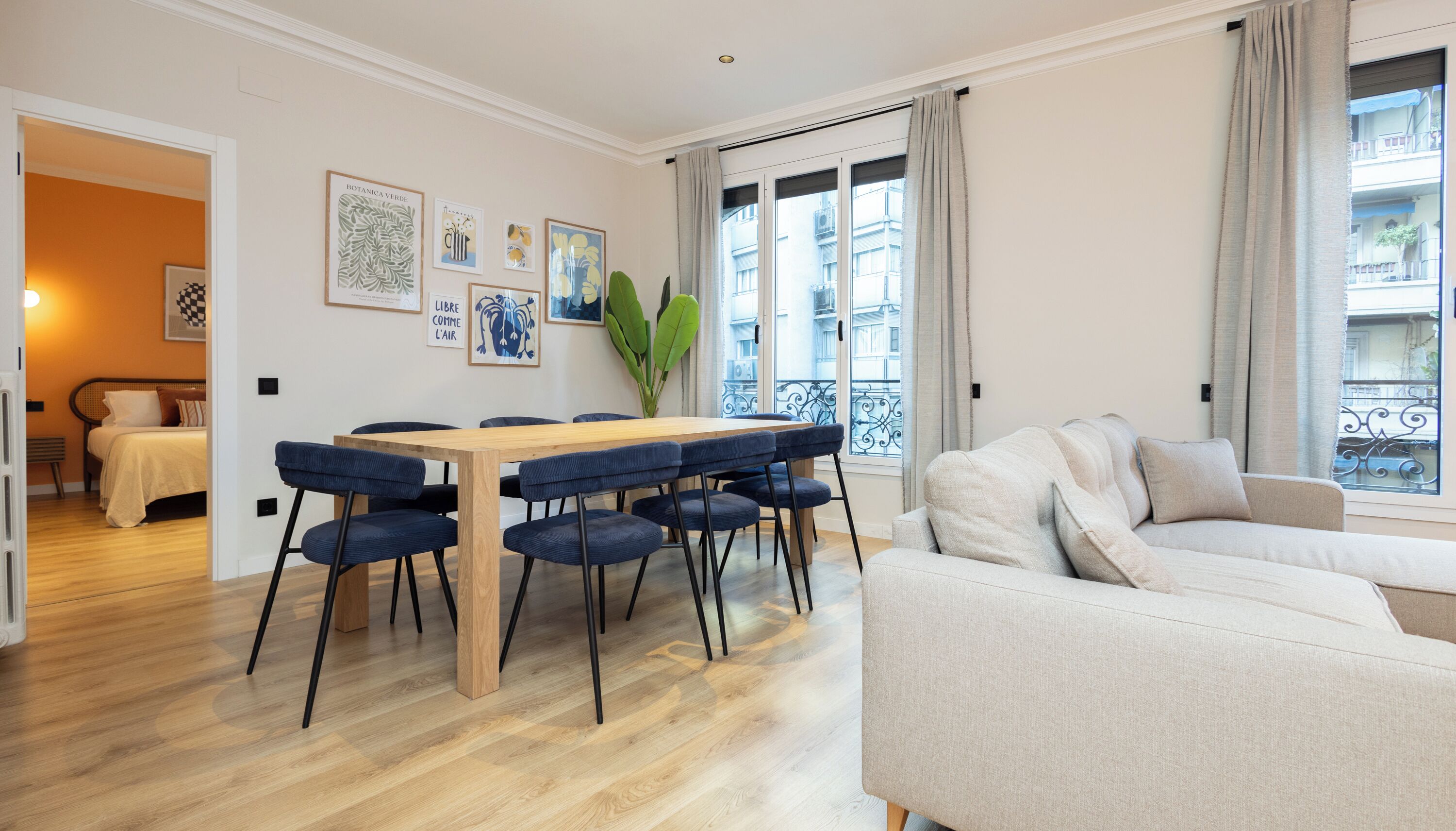 Un salon moderne avec une table à manger en bois et des chaises bleues, un canapé beige et une grande fenêtre donnant sur des immeubles.