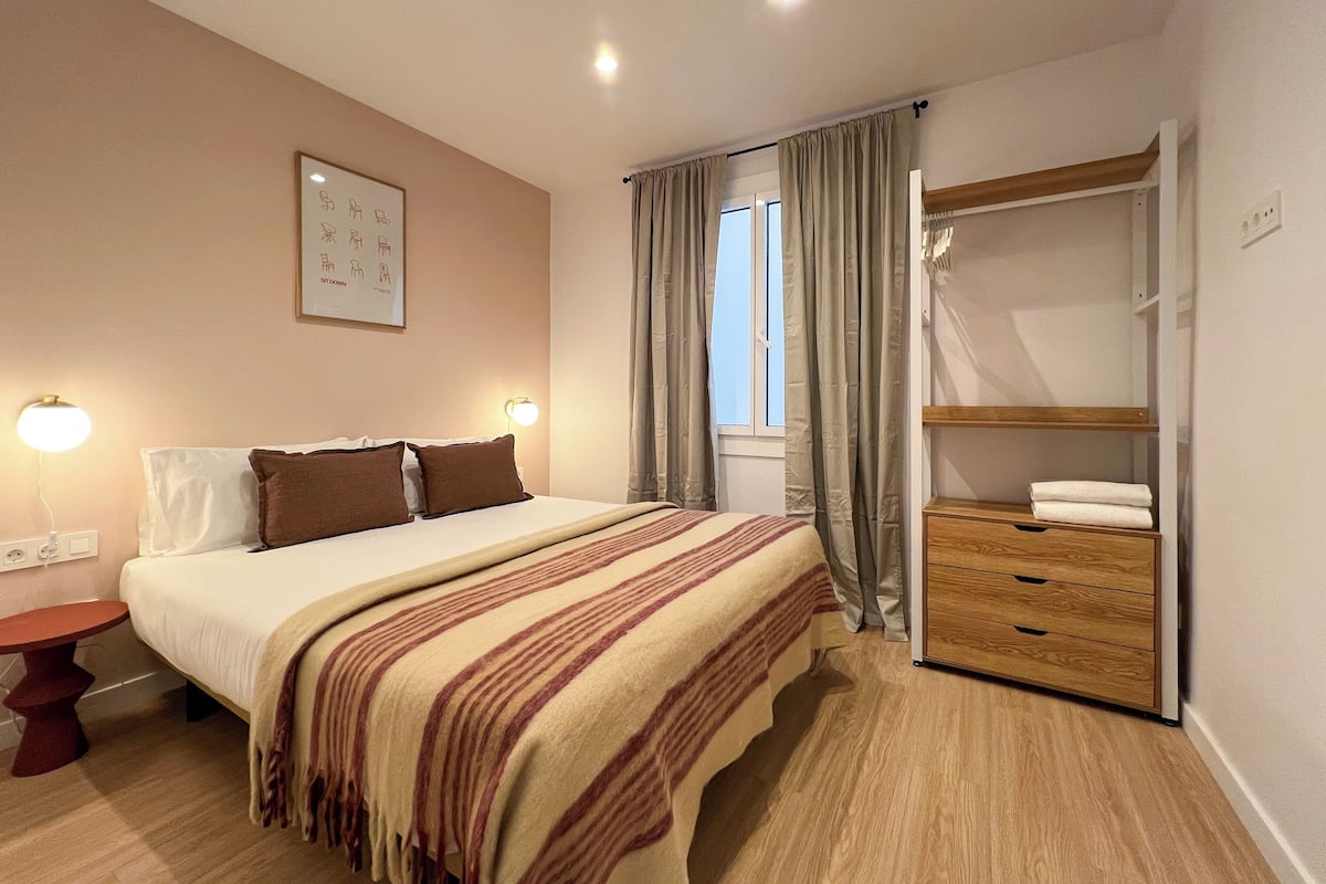 Ein Schlafzimmer mit einem Bett, Nachttischlampen, einem Fenster mit Vorhängen, einer Holzkommode und einem gerahmten Bild an der Wand.