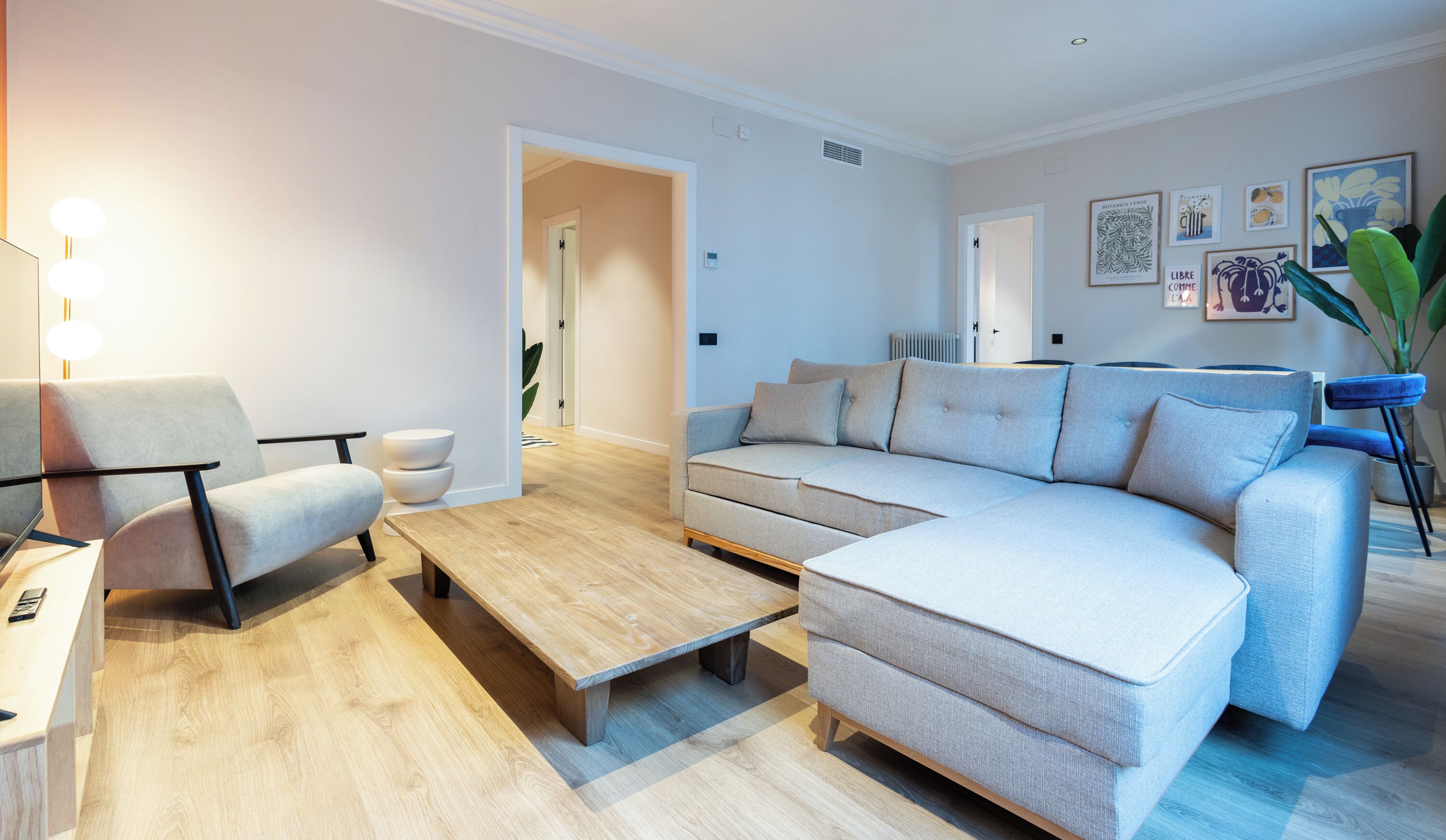 Un salon moderne avec un canapé gris, une table basse en bois et un fauteuil blanc. On aperçoit une plante dans un coin et des œuvres d’art accrochées au mur.