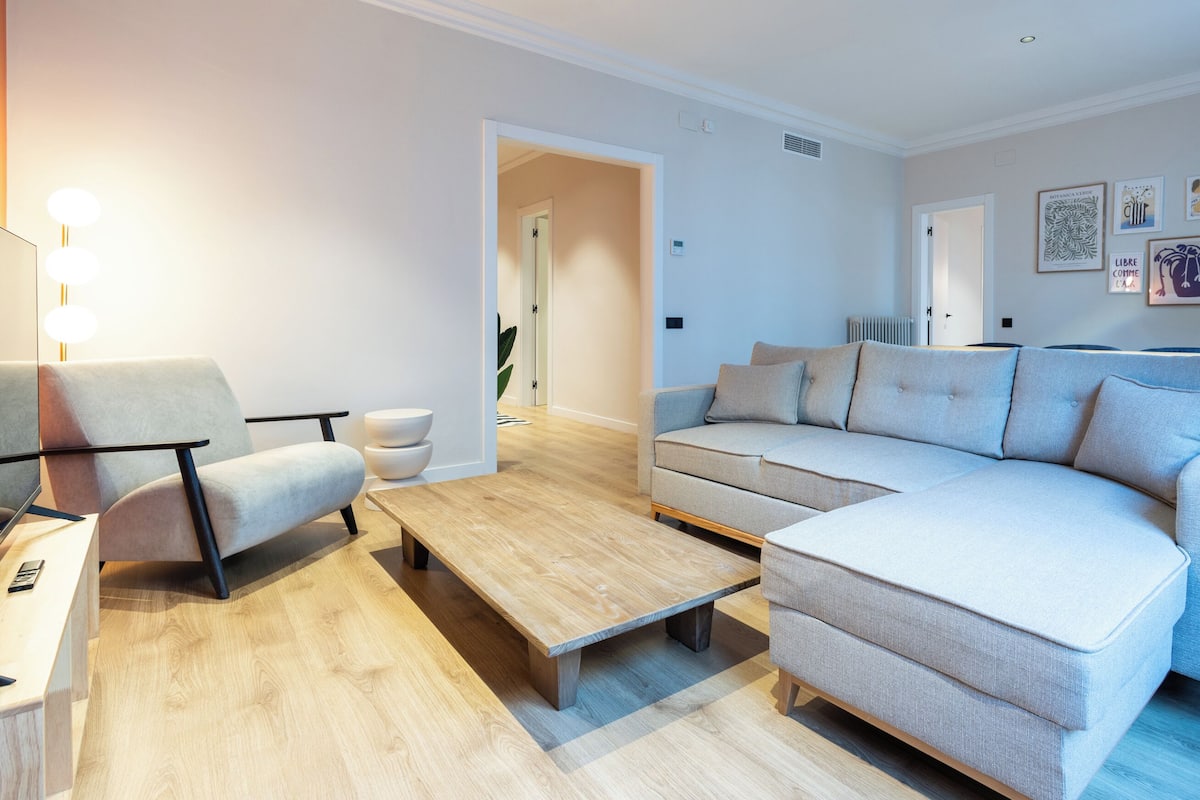 Ein modernes Wohnzimmer mit einem grauen Sofa, einem Holztisch und einem weißen Sessel. In der Ecke steht eine Pflanze und an der Wand hängt ein Kunstwerk.
