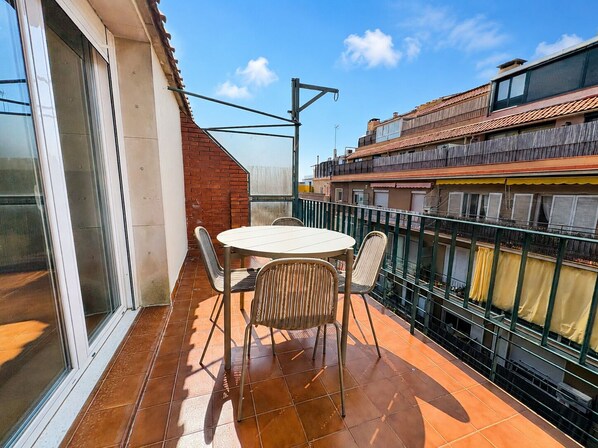 Un balcón con una mesa redonda y sillas, vista de edificios cercanos y cielo despejado.