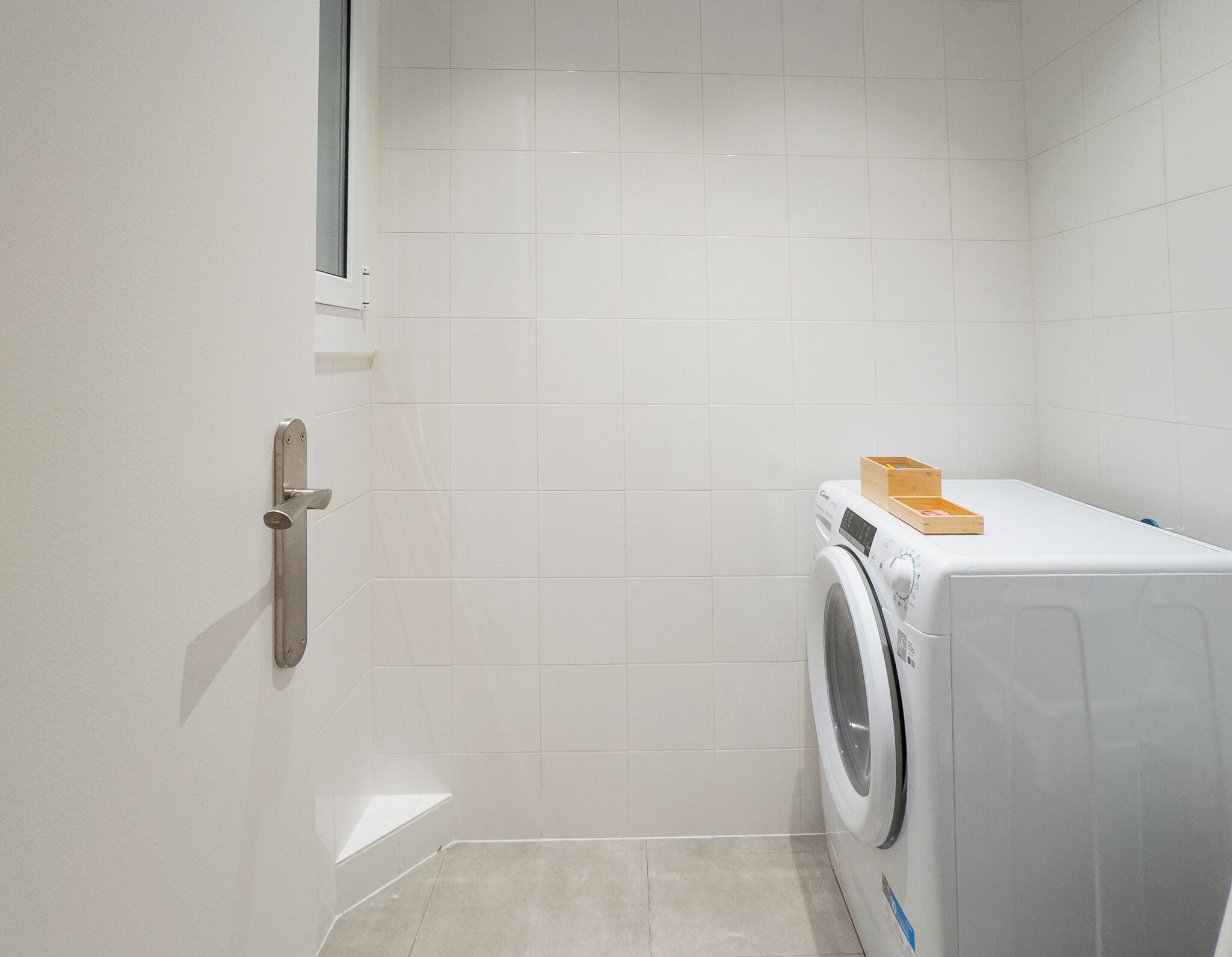 A compact, modern laundry room with a front-loading washing machine, a wooden box on top, and a door with a handle.