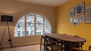 Ein Essbereich mit einem runden Holztisch, Korbstühlen, einer Stehlampe und einem großen, rundbogigen Fenster mit aufwendiger Eisenverzierung.