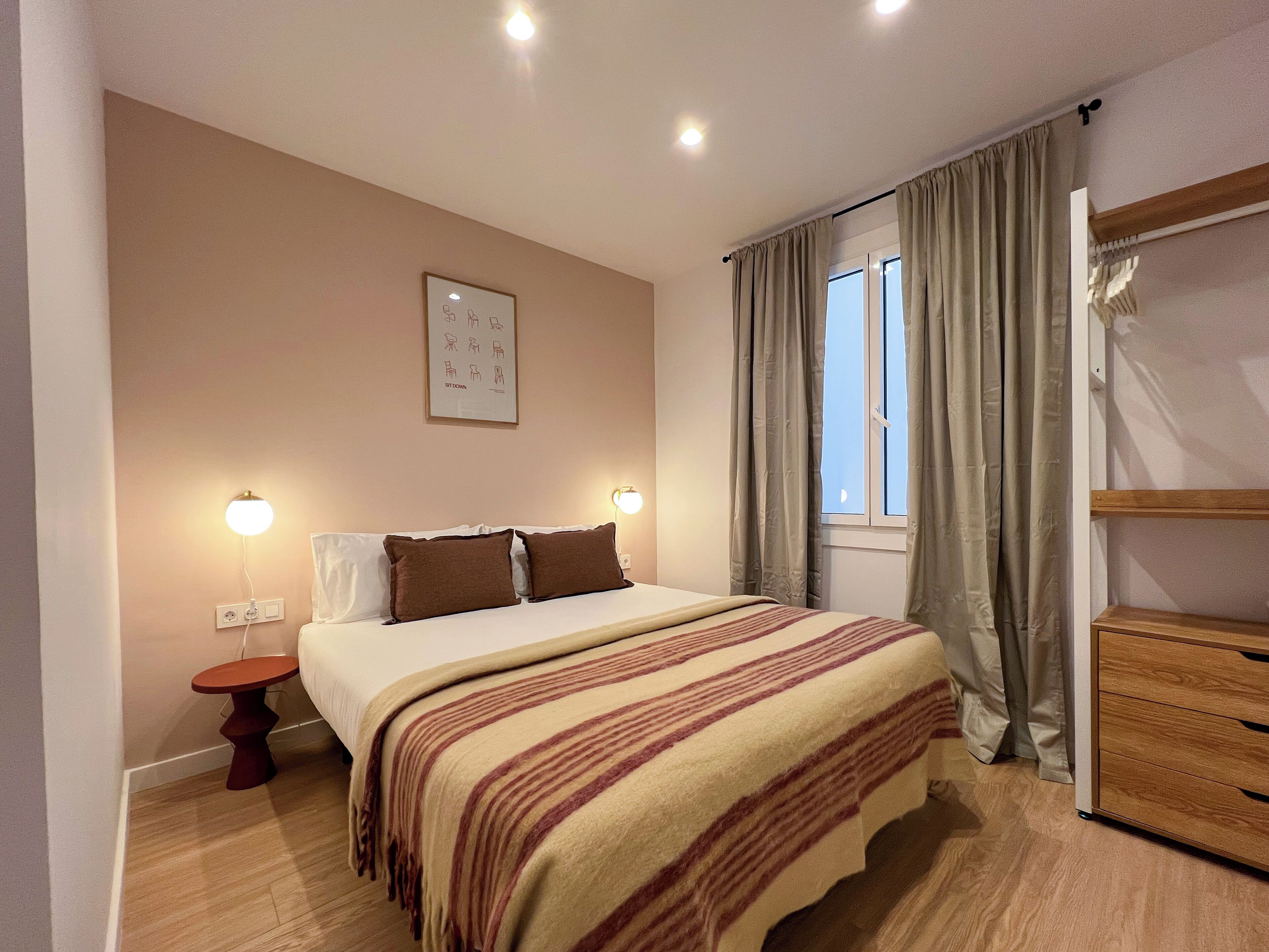 Een slaapkamer met een bed, nachtlampjes, een raam met gordijnen, een houten dressoir en een wandlamp.