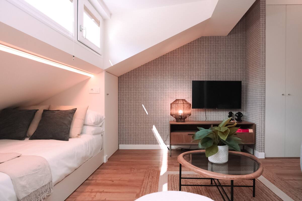 A modern living room with a sloped ceiling, a flat-screen TV mounted on the wall, a wooden coffee table with a plant, and a sofa with a throw blanket.