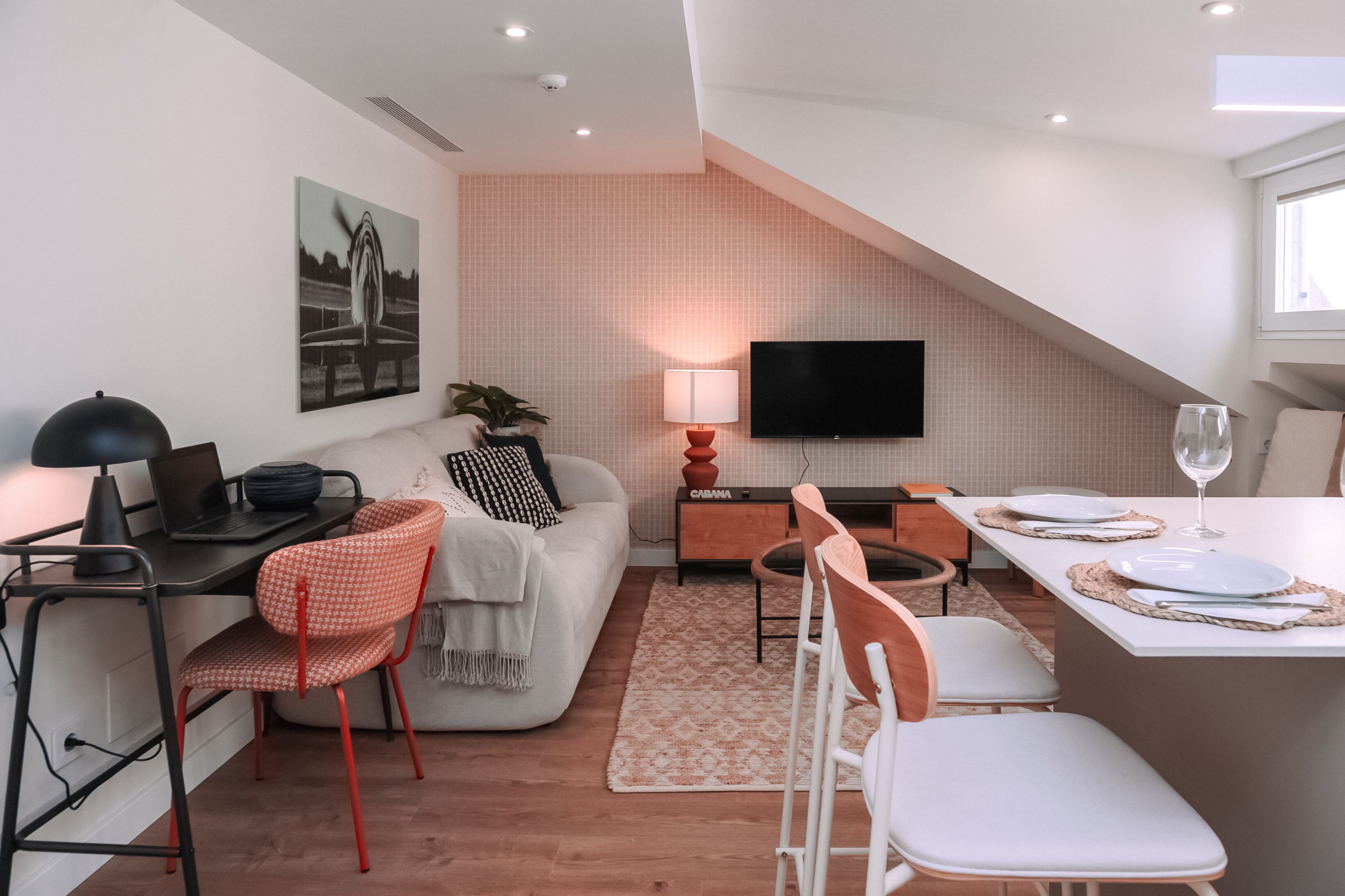 Una sala moderna con sofá, una mesa de comedor para dos personas, televisor montado en la pared y una lámpara.