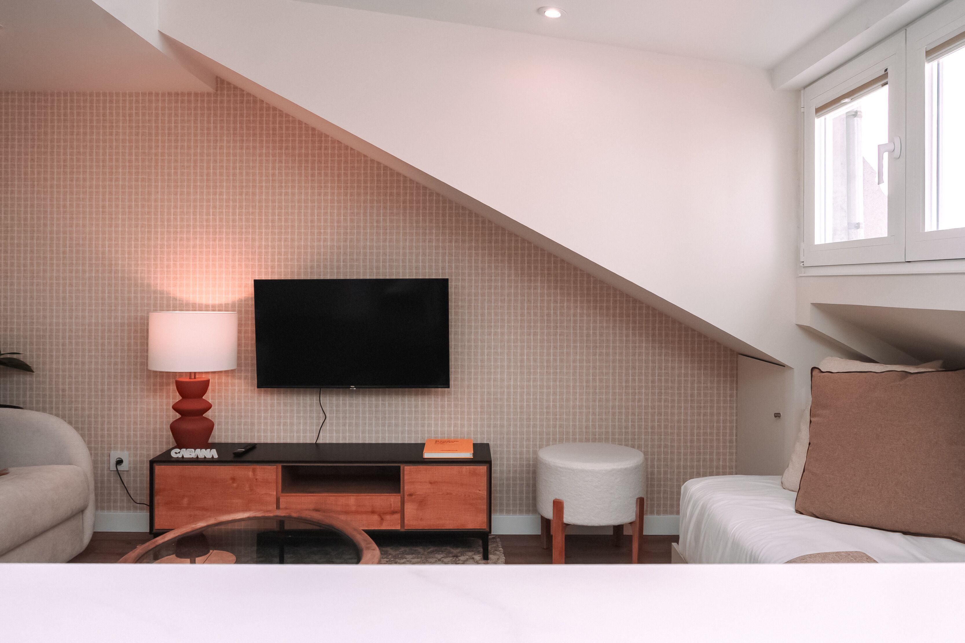 A modern hotel room with a flat-screen TV mounted on the wall, a wooden console table, a round white ottoman, a bed with a large pillow, and a window with white shutters.