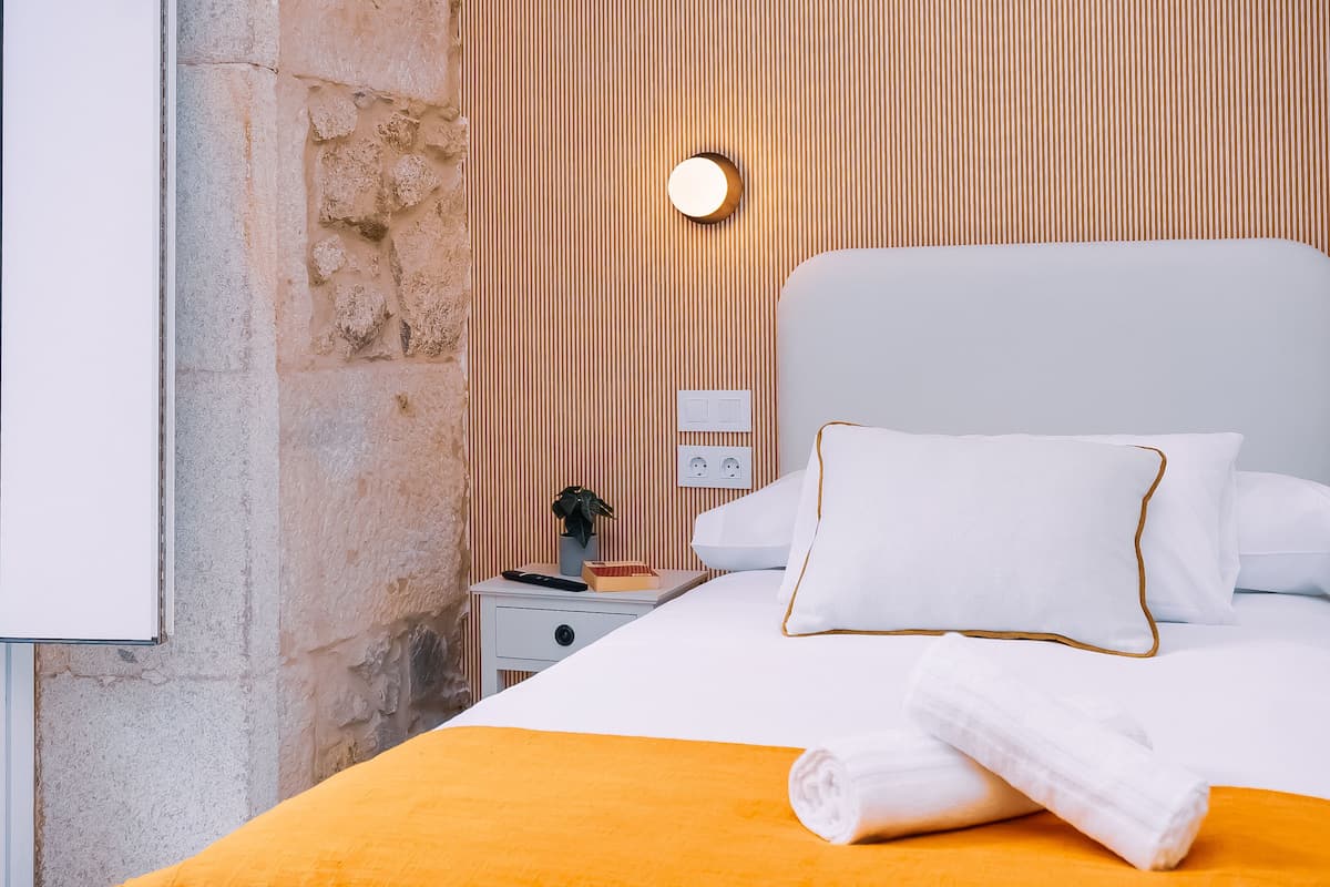 A neatly made bed with white bedding and a yellow throw, a bedside table with a lamp, and a wall-mounted light fixture.