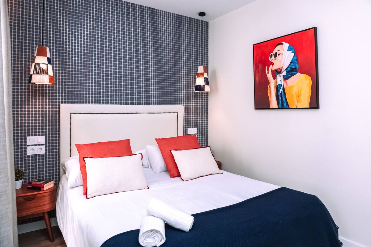 A neatly made bed with white and red pillows, a blue blanket, and a rolled towel. There are two wall-mounted lamps and a painting on the wall.