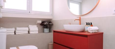 Un baño moderno con inodoro blanco, tocador rojo y toallas ordenadas en un estante.