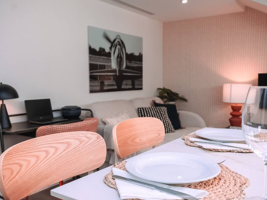 A dining area with a white table set for two, a laptop on a desk, a sofa with cushions, and a painting on the wall.