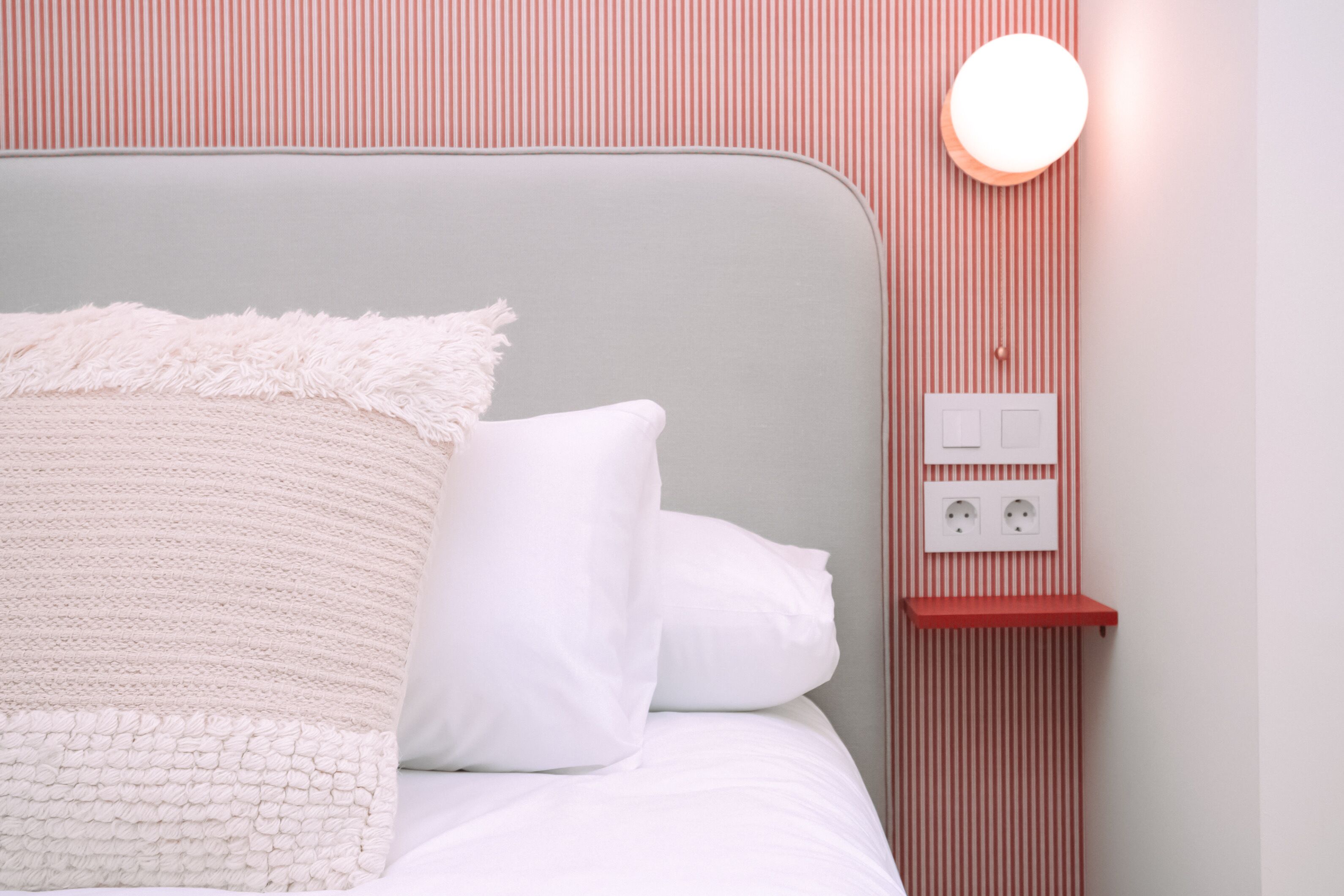 A neatly made bed with white bedding and a fluffy blanket, a bedside table with a red shelf, and a wall-mounted light fixture.