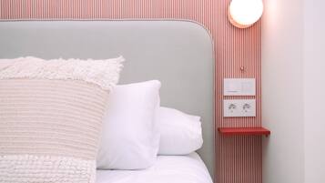 A neatly made bed with white bedding and a fluffy blanket, a bedside table with a red shelf, and a wall-mounted light fixture.