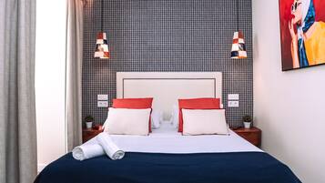 A neatly made bed with white and red pillows, a blue bedspread, and a rolled towel on the side. There are two bedside tables with lamps and a small potted plant on each. The wall behind the bed has a checkered pattern.