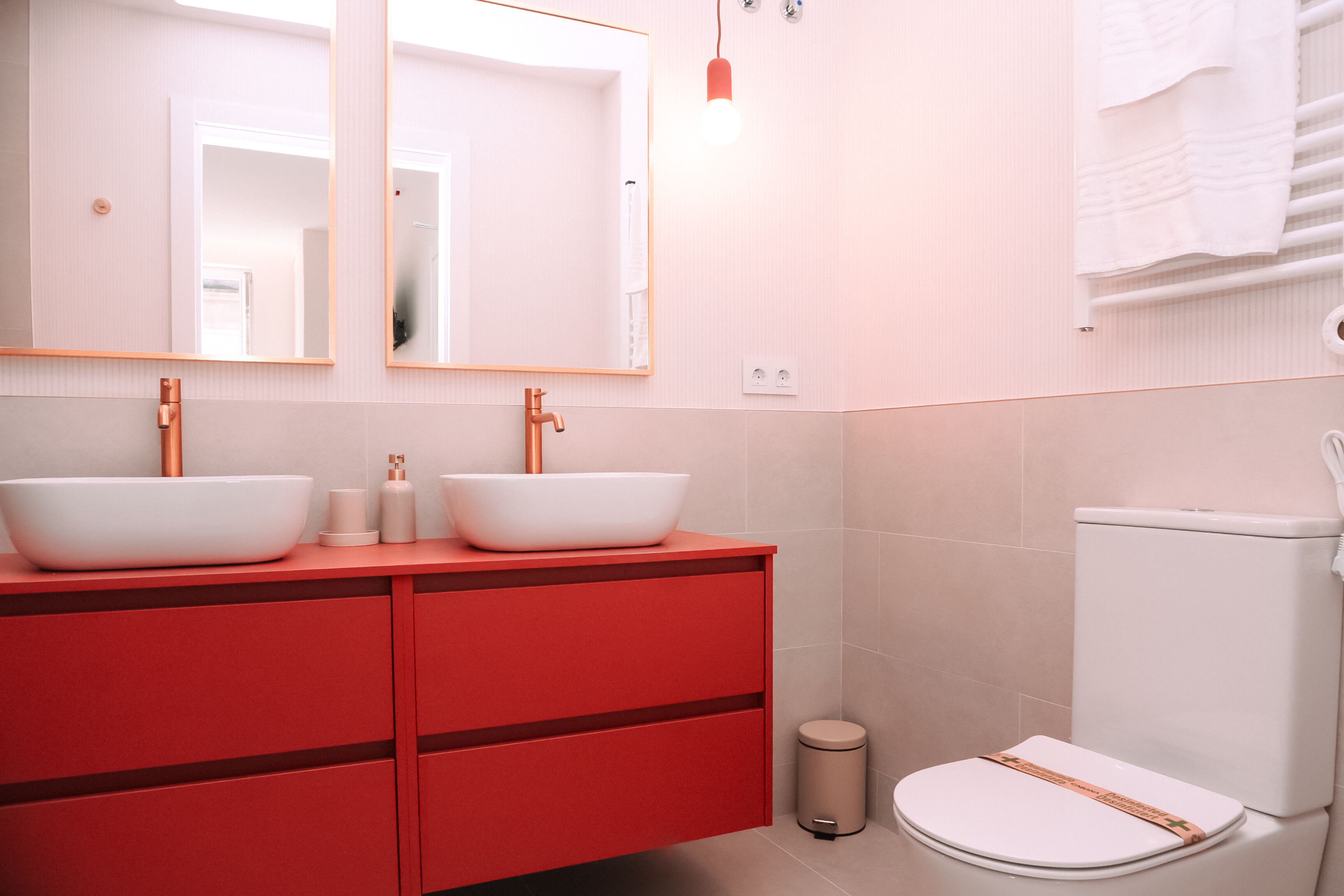 Et badeværelse med to hvide håndvaske i skåle, et rødt kommodebord, et hvidt toilet og en lille skraldespand.