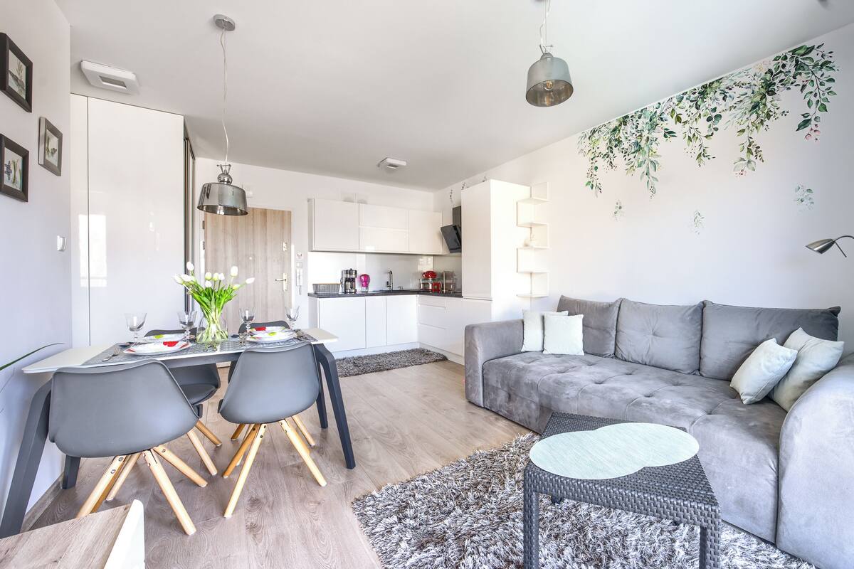 Ein modernes Wohnzimmer mit einem grauen Sofa, einem Esstisch mit Stühlen und einer Küche im Hintergrund.