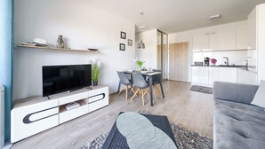 Ein modernes Wohnzimmer mit Flachbildfernseher, einem Esstisch mit Stühlen, einem grauen Sofa und einer Küchenzeile mit weißen Schränken.