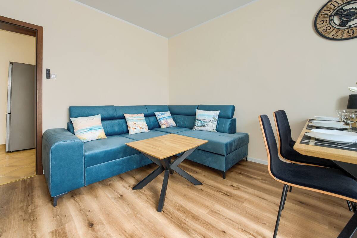 Ein modernes Wohnzimmer mit einem blauen Sofa, einem Holztisch und einem Essbereich, gedeckt für zwei Personen.