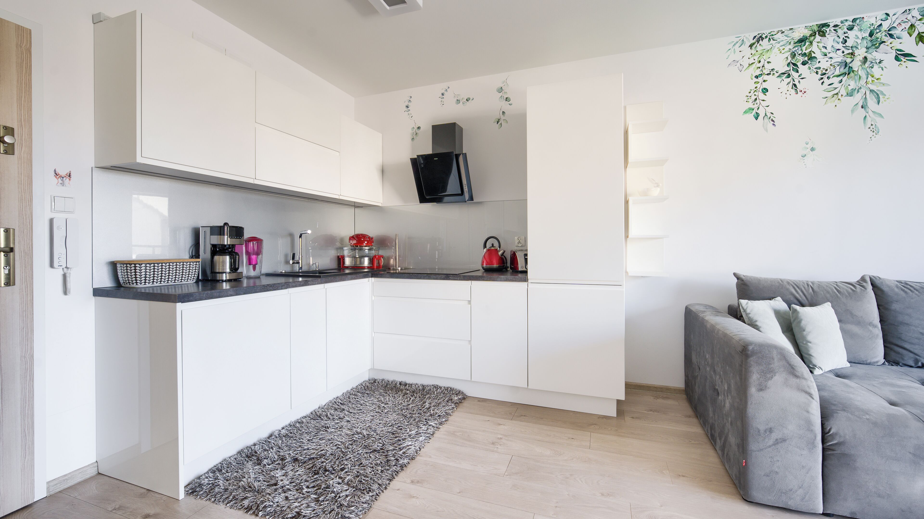 Une cuisine moderne avec des meubles blancs, un réfrigérateur noir et un canapé gris dans l’espace salon.