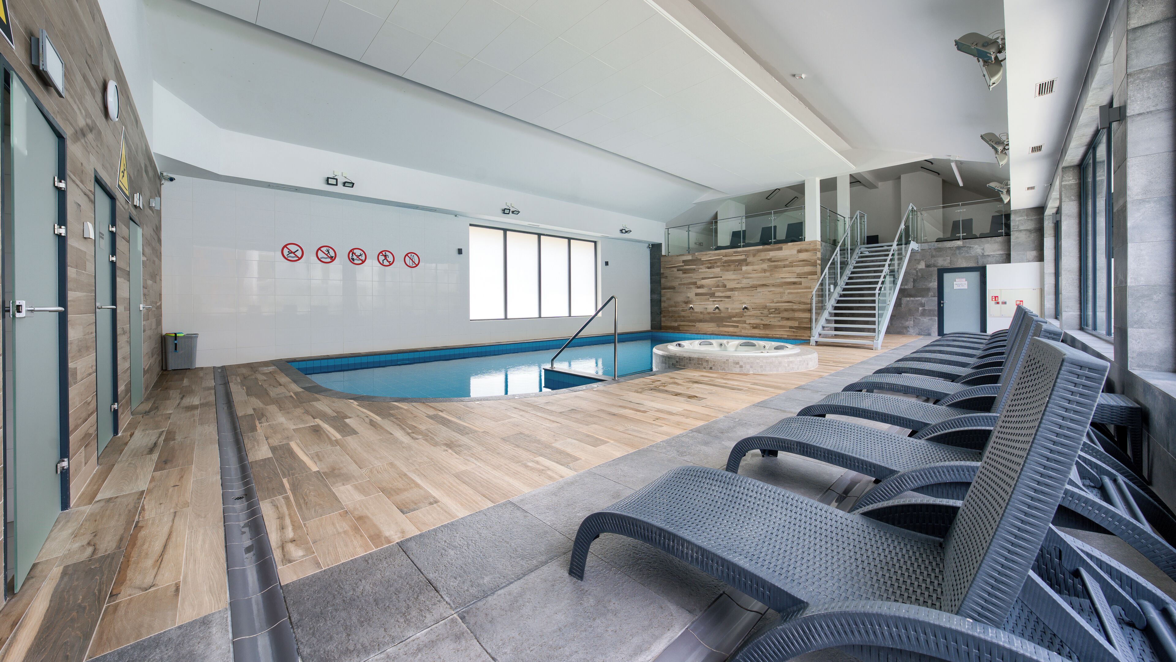 A modern indoor swimming pool with a wooden floor, a hot tub, and lounge chairs.