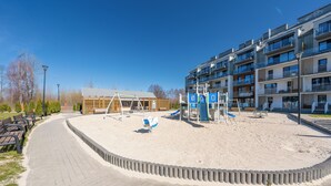 Ein Spielplatz mit Sand, Schaukeln und einer Rutsche, umgeben von einem Gebäude mit mehreren Balkonen und einem gepflasterten Weg.