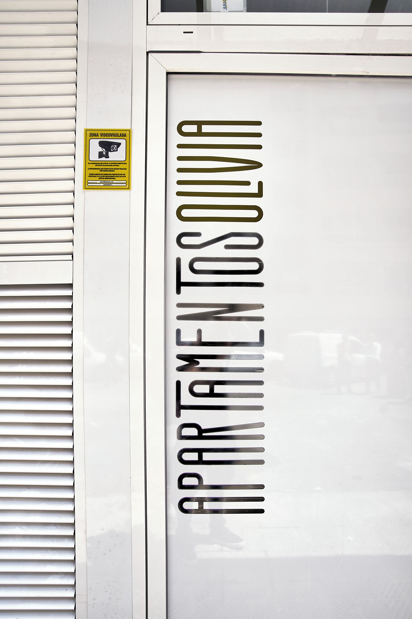 Eine weiße Tür mit dem Schriftzug "APARTAMENTOSOLUVIOLA" in fetten, schwarzen Buchstaben. Auf der Tür ist ein gelbes Schild mit einem Symbol für eine Überwachungskamera und Text zu sehen.