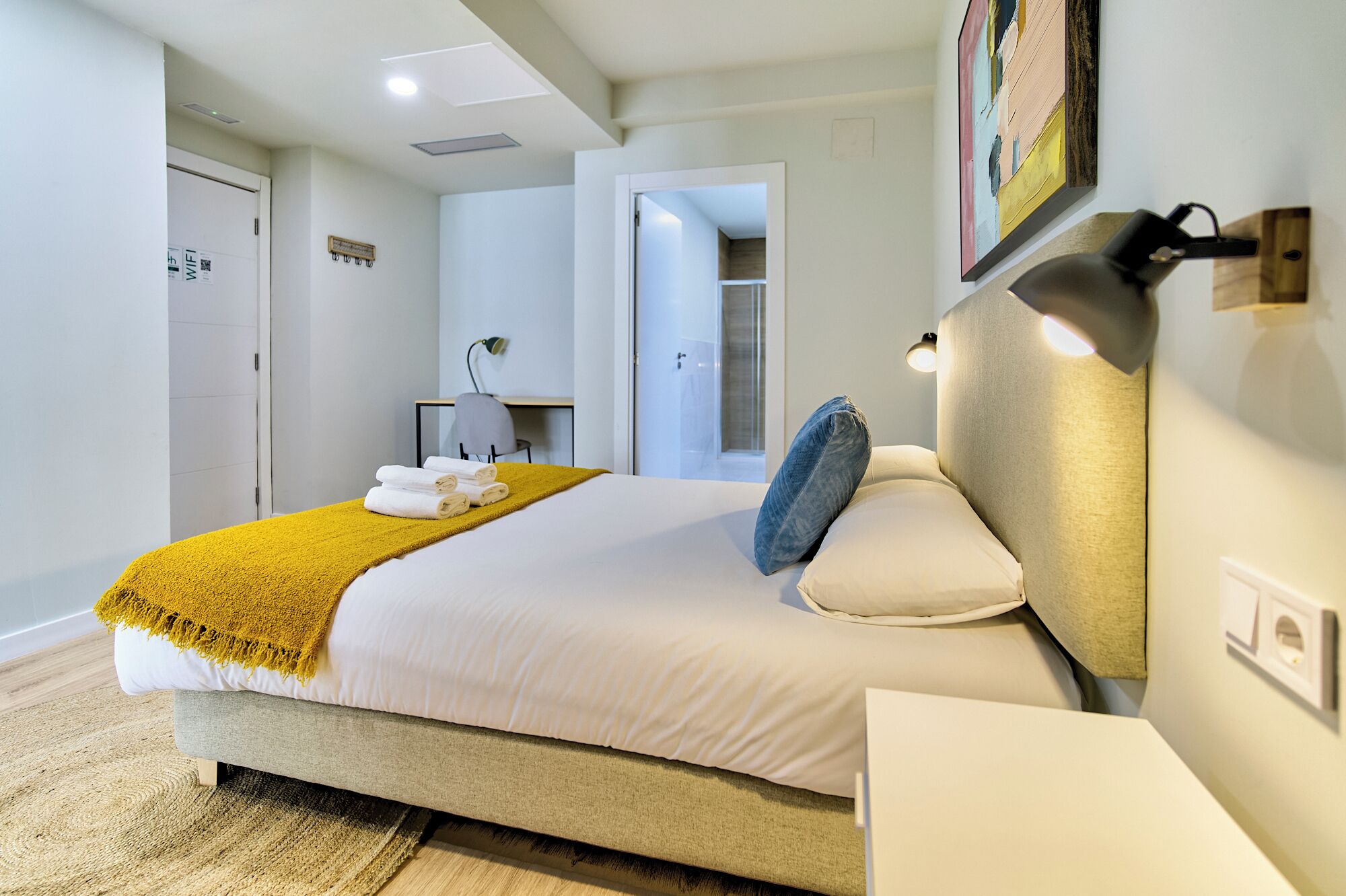 A modern hotel room with a bed, a desk, a chair, and a bathroom visible through an open door.