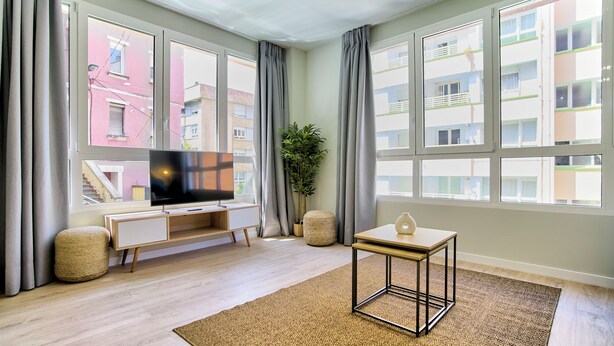 Ein modernes Wohnzimmer mit Flachbildfernseher, einem kleinen Tisch und einem großen Fenster mit Blick auf Gebäude.