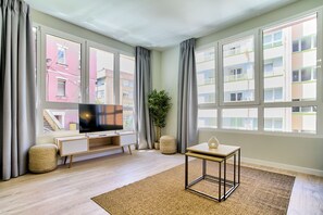 Una sala moderna con un televisor de pantalla plana, una mesita pequeña y un ventanal con vistas a edificios.