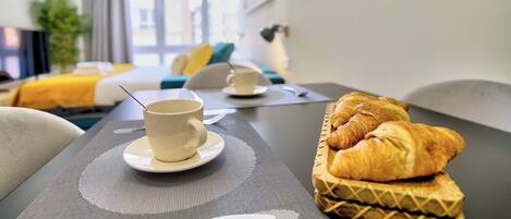 Une chambre d’hôtel avec une table à manger dressée, agrémentée d’une tasse et de croissants, un lit avec une literie jaune et une vue sur la ville par de grandes fenêtres.
