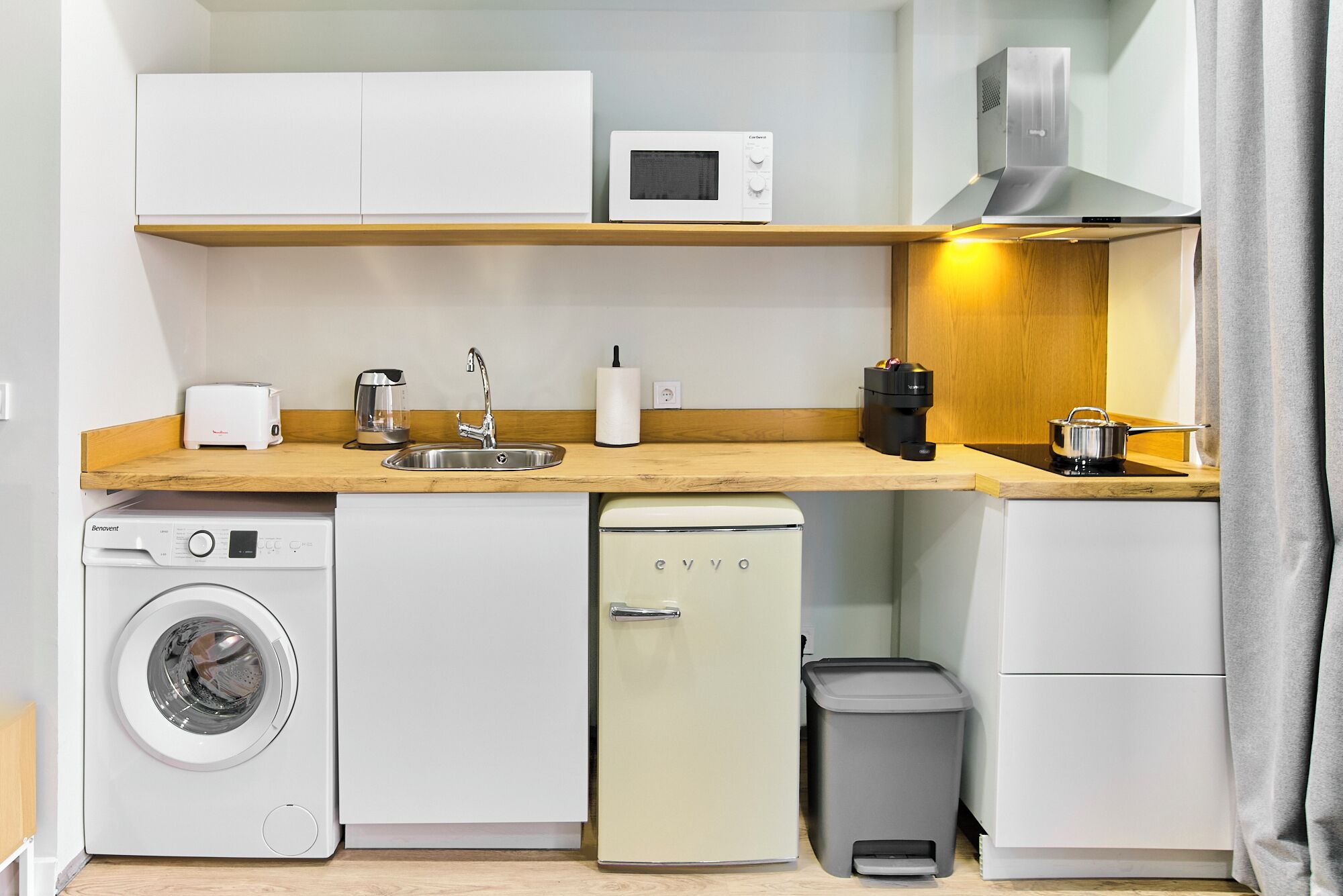 A compact kitchenette with a washing machine, a small refrigerator, a microwave, a sink, and a stove.
