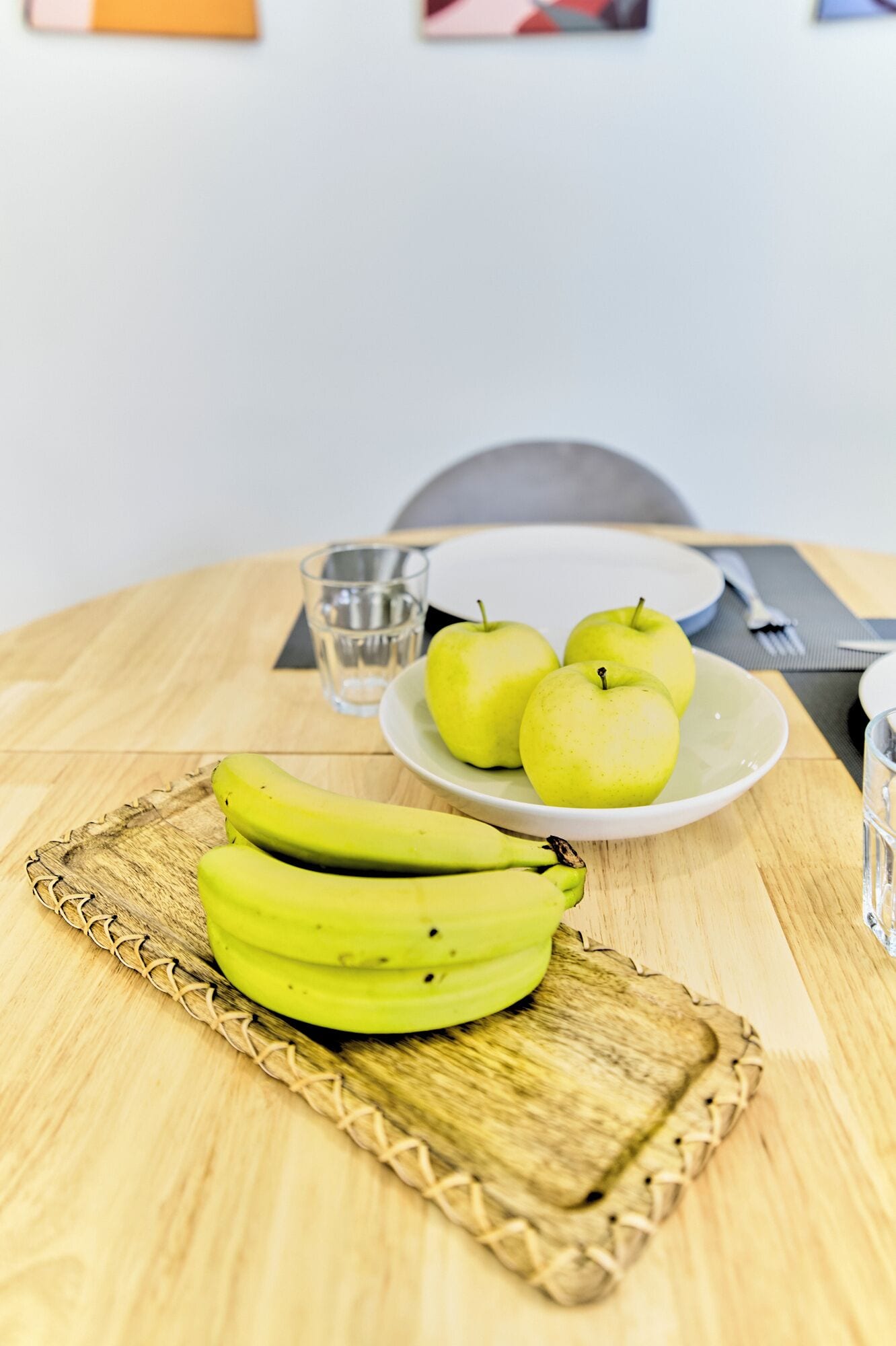 A wooden table with a woven placemat, a plate of green apples, a glass of water, and a banana bunch.