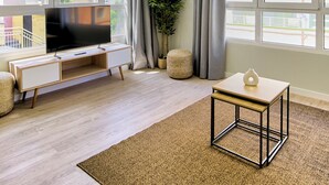 Ein modernes Wohnzimmer mit Flachbildfernseher, einem kleinen Holztisch und einem großen Fenster mit Vorhängen.