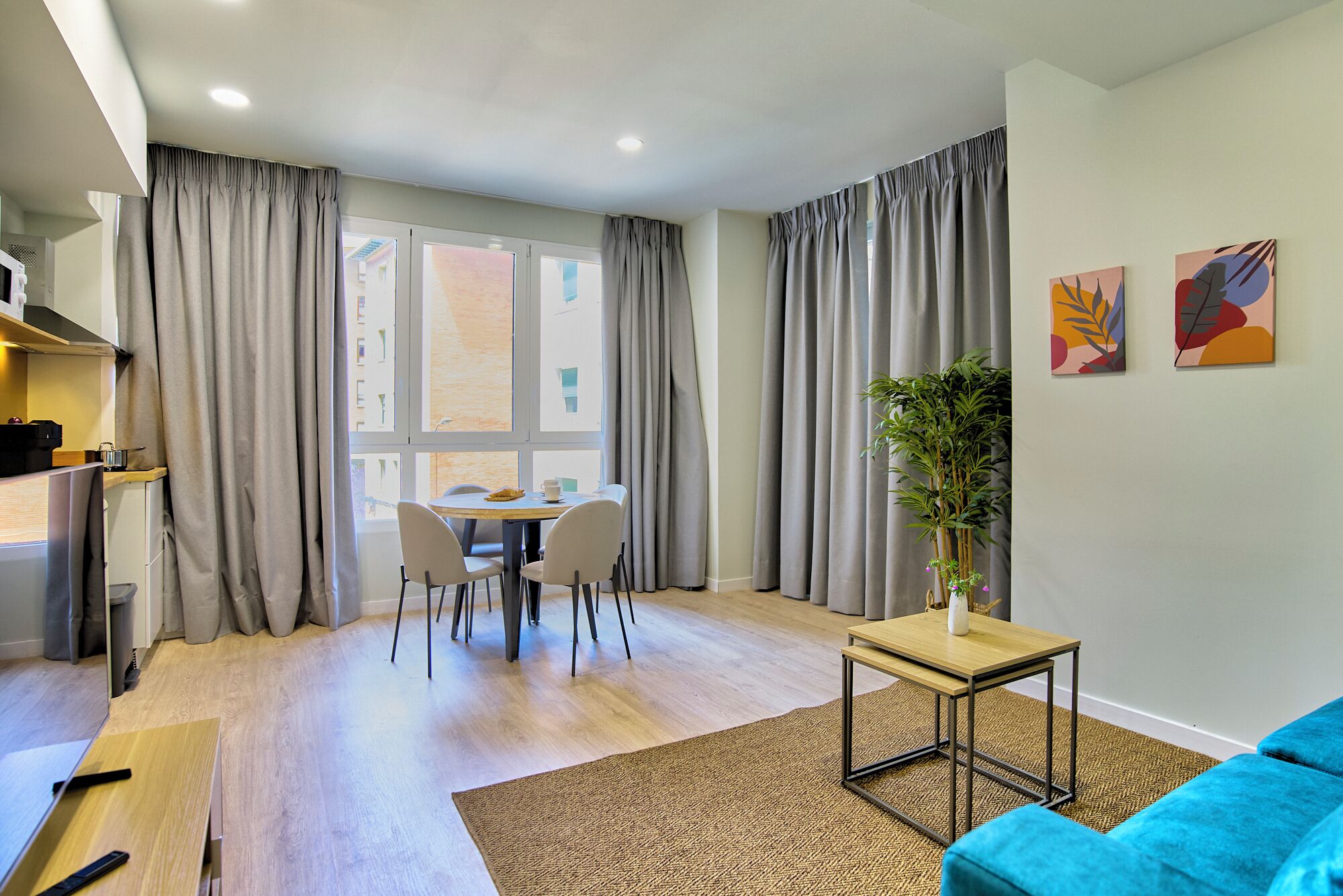 Een moderne woonkamer met een eethoek, grote ramen voorzien van gordijnen, een blauwe bank en twee abstracte schilderijen aan de muur.
