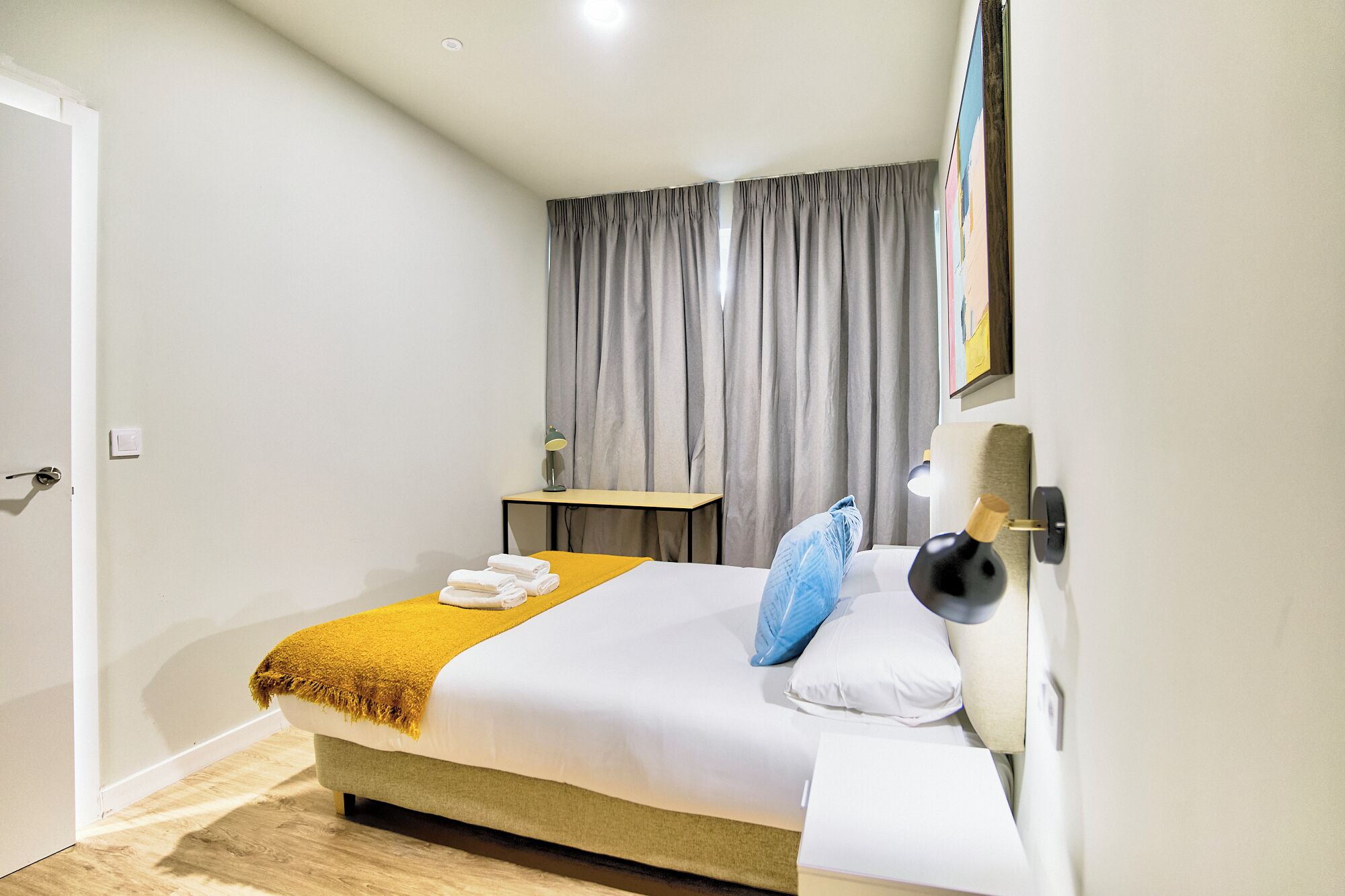 A modern hotel room with a large bed, a bedside table, a lamp, and a painting on the wall.