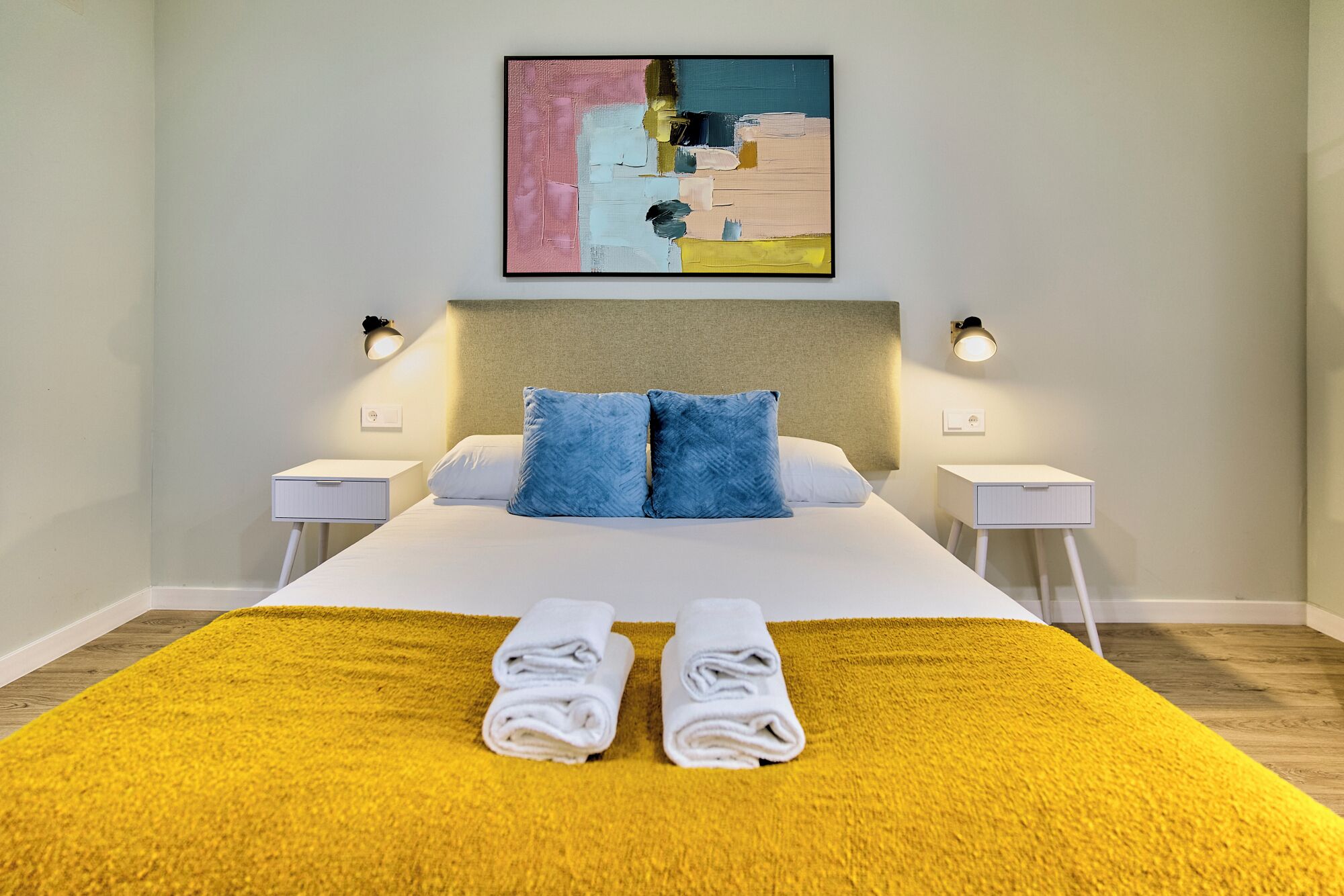 Une chambre d’hôtel avec un lit, deux tables de chevet, une tête de lit, un tableau et deux lampes.
