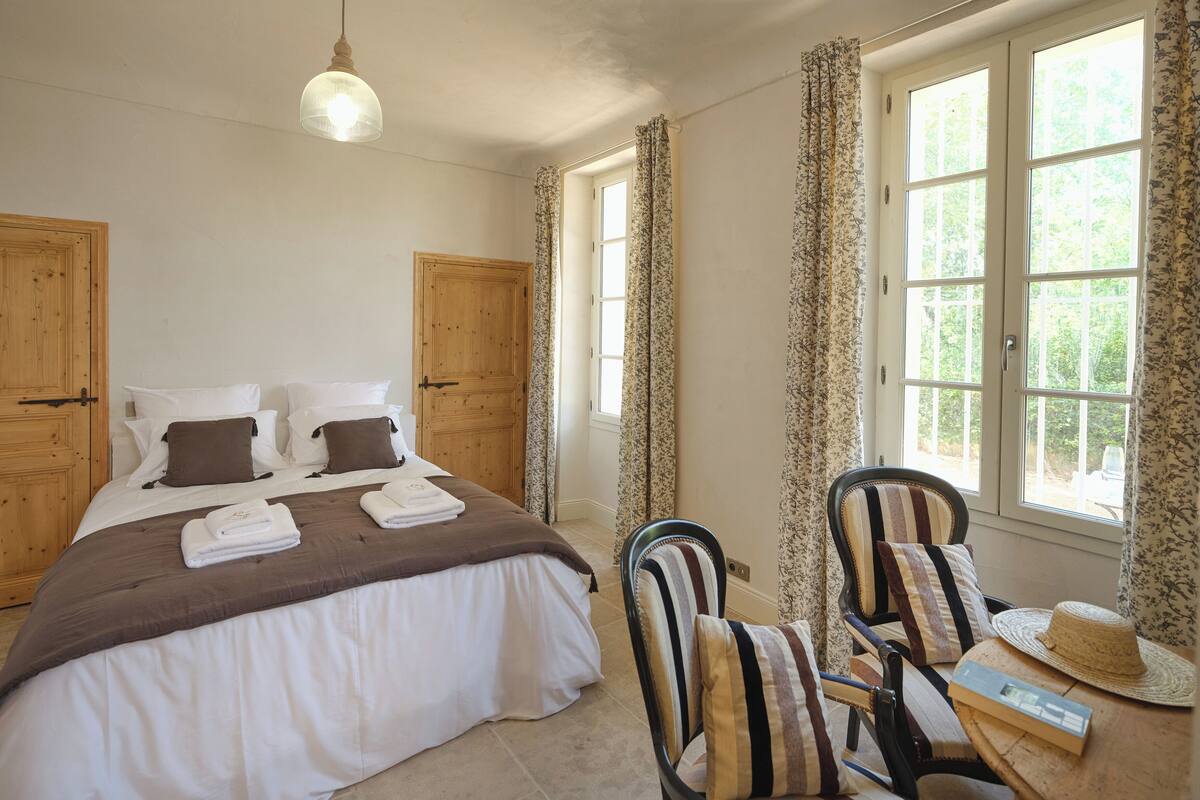 A bedroom with a bed, two chairs, a hat, and a book on a table.