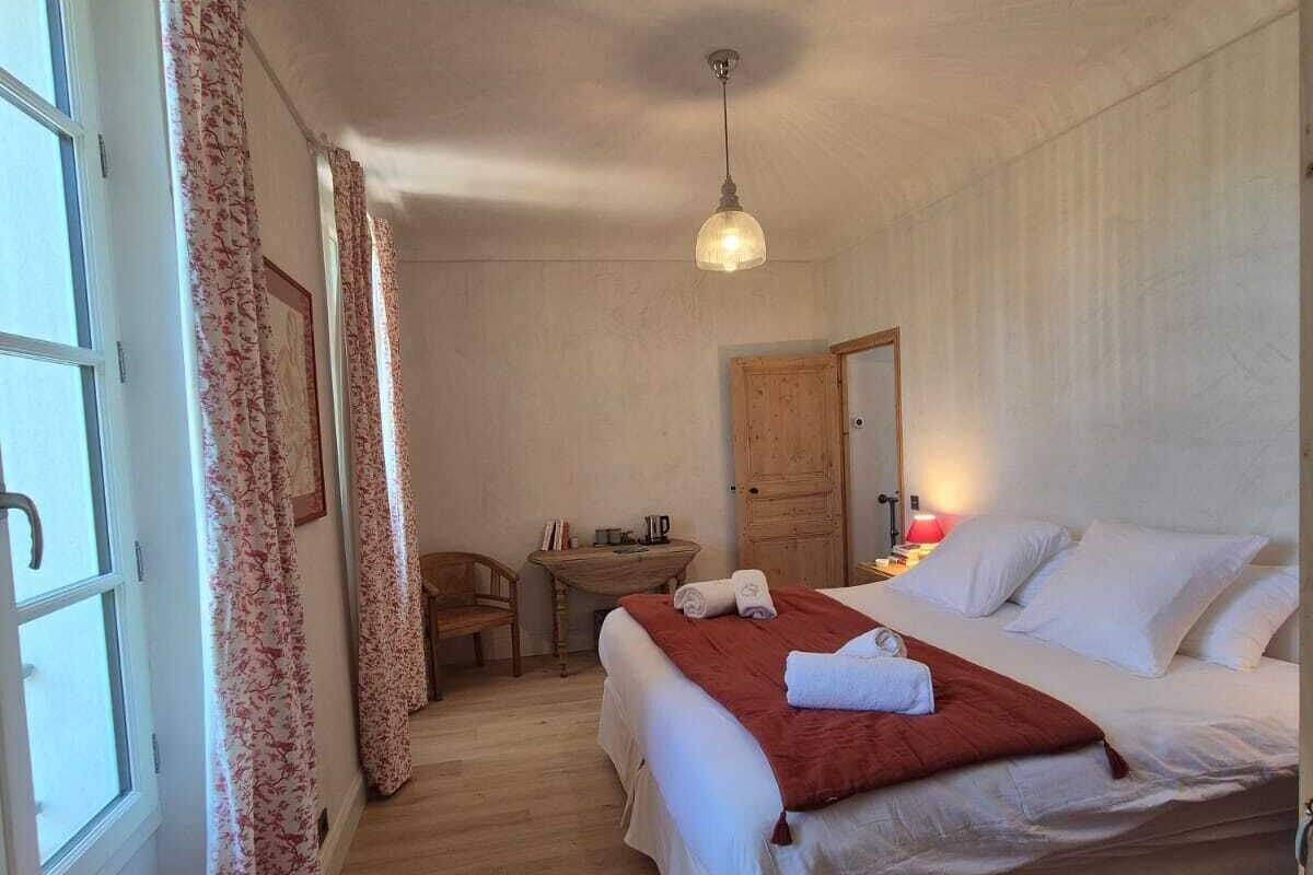 A bedroom with a large bed, a wooden desk, a chair, a small table, and a window with curtains.