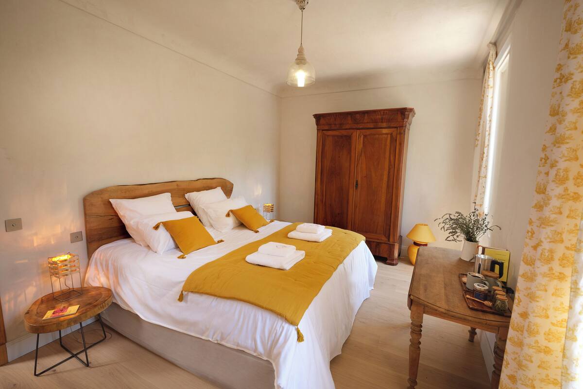A neatly made bed with white and yellow bedding, a wooden headboard, a bedside table with a lamp, and a wardrobe in a well-lit room.