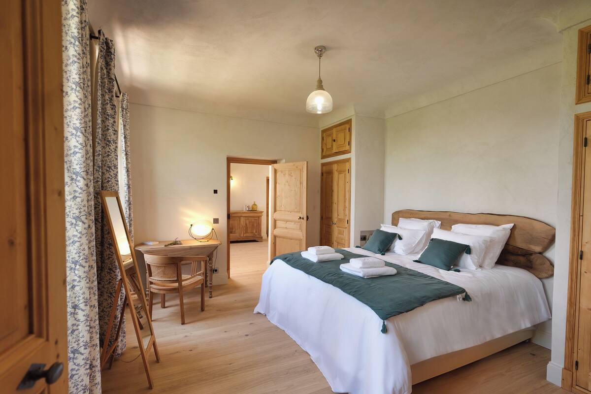 A bedroom with a wooden bed, white bedding, a desk with a lamp, and a wooden floor.